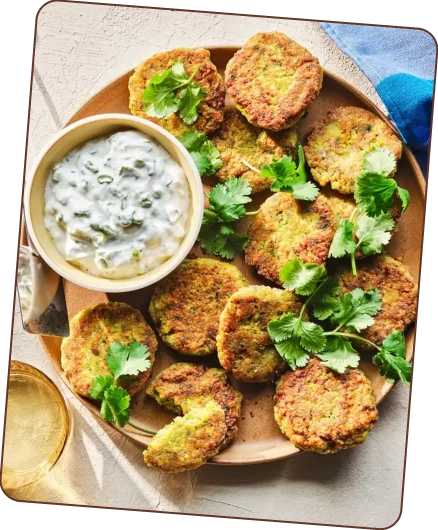 A plate of golden-brown fritters, likely falafel or crab cakes, garnished with fresh cilantro and served with a side of white dipping sauce.