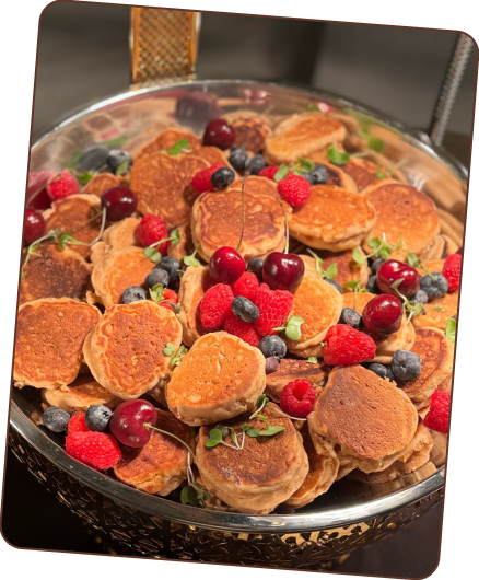 A large serving dish filled with mini pancakes, topped with fresh raspberries, blueberries, and cherries.